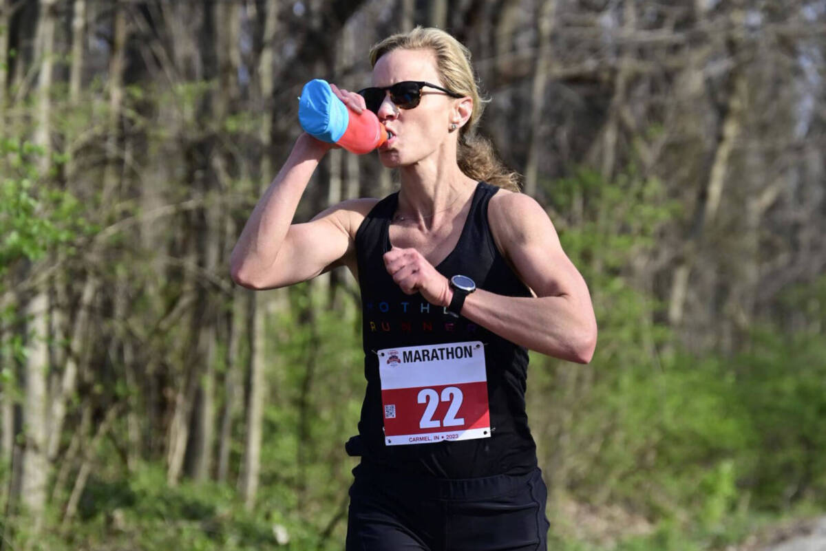 Whitney Heins running a marathon and drinking from a handheld water bottle.