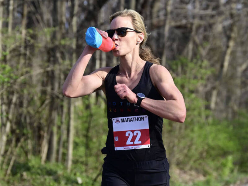 Whitney Heins running a marathon and drinking from a handheld water bottle.