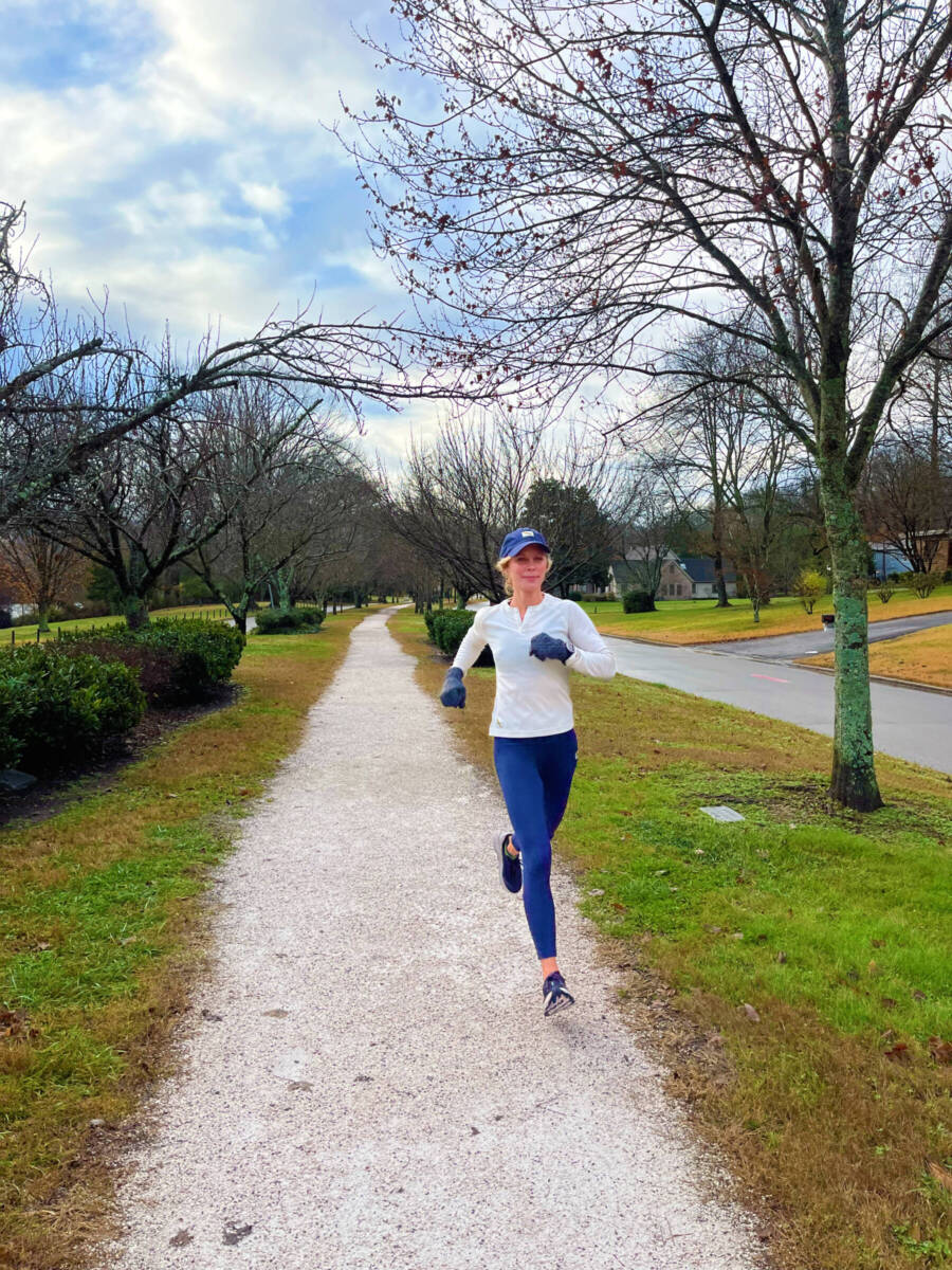 Whitney Heins running in a white shirt and blue tights and blue hat on gravel path. 