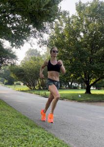 Woman running in orange shoes, black sports bra, and green shorts. 